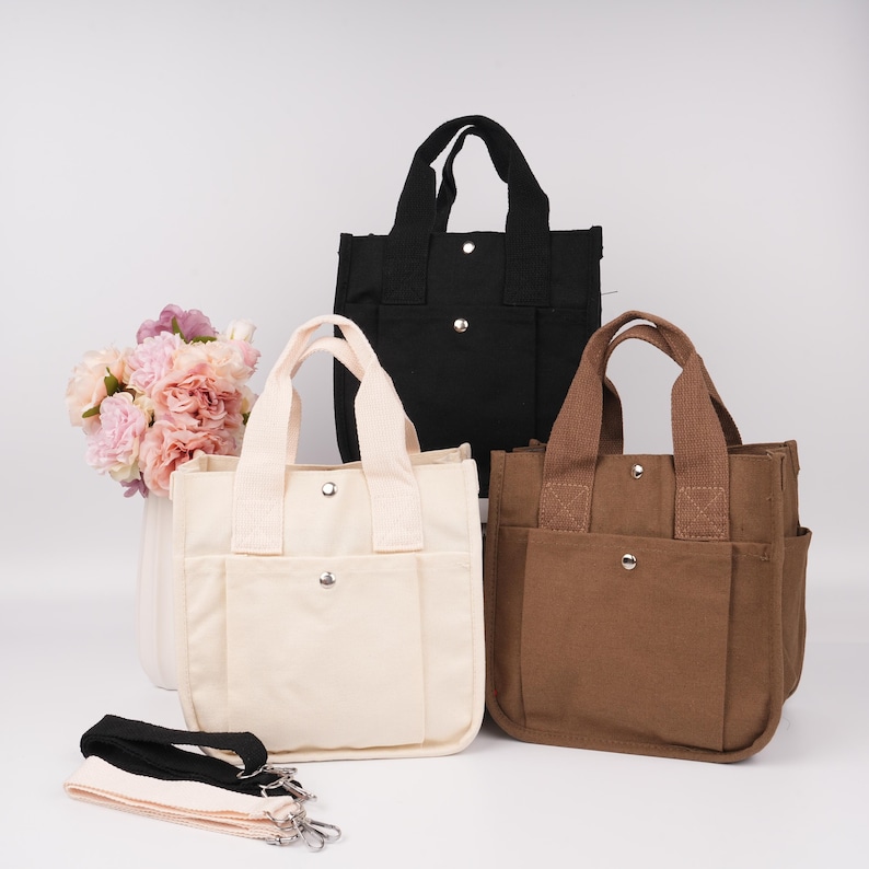 Pode incluir: Tr&ecirc;s sacolas de lona nas cores preta, creme e marrom, cada uma com duas al&ccedil;as superiores, um bolso frontal com fecho de bot&atilde;o de press&atilde;o prateado e uma al&ccedil;a de ombro remov&iacute;vel. As bolsas est&atilde;o dispostas com um buqu&ecirc; de flores rosas.