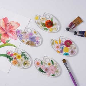 May include: Five clear resin palettes shaped like artist palettes, each embedded with various pressed dried flowers and green foliage. Two paint tubes and a paintbrush are positioned nearby, alongside a sheet of paper with a floral watercolor painting.