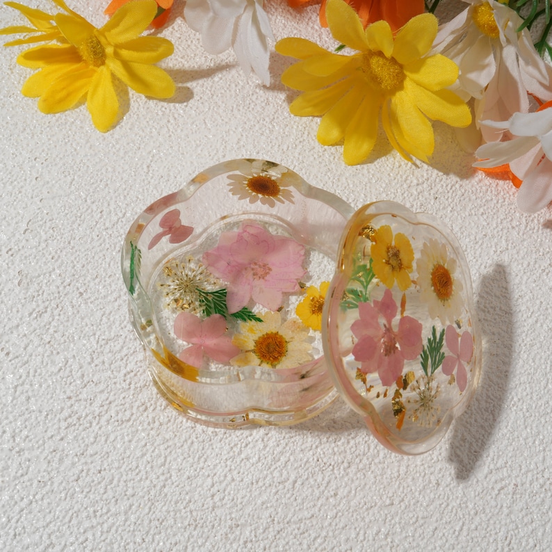 K&ouml;nnte beinhalten: Eine transparente Schmuckschatulle aus Harz mit einem gewellten Rand und einem Deckel. Die Schachtel und der Deckel sind mit gepressten Blumen in Rosa-, Gelb- und Wei&szlig;t&ouml;nen verziert.