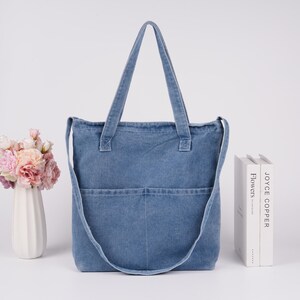 Peut inclure: Sac fourre-tout en jean bleu clair avec deux poches avant et une longue bandouli&egrave;re. Le sac a deux poign&eacute;es courtes. Un vase de fleurs et deux livres sont en arri&egrave;re-plan. Le sac est sur une surface blanche.