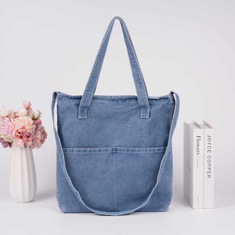 Peut inclure: Sac fourre-tout en jean bleu clair avec deux poches avant et une longue bandouli&egrave;re. Le sac a deux poign&eacute;es courtes. Un vase de fleurs et deux livres sont en arri&egrave;re-plan. Le sac est sur une surface blanche.