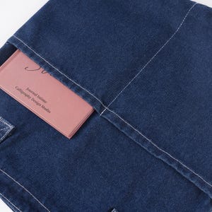 Peut inclure: Gros plan sur une couverture de journal en denim bleu fonc&eacute; avec des coutures blanches. Un journal rose est partiellement visible dans la poche, avec le texte "Journal Intime Calligraphy Design Studio".