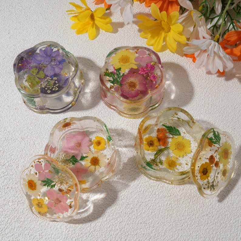 K&ouml;nnte beinhalten: Vier transparente Schmuckk&auml;stchen aus Harz mit Blumenakzenten. Jede Box zeigt eine andere Anordnung von gepressten Blumen in den Farben Rosa, Lila, Gelb und Orange.