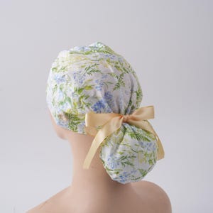 May include: A scrub cap featuring a white fabric base with a botanical print of green leaves and small blue and yellow flowers. The cap includes a light beige satin ribbon tied into a bow at the back and a small button on the side for securing masks.