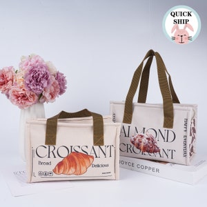 May include: Two beige insulated lunch bags with brown handles. One bag features a croissant with the text "Croissant, Bread, Delicious". The other bag features an almond croissant with the text "Almond Croissant, Joyce Copper, Happy Everyday".