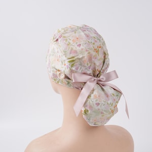 May include: A scrub cap featuring a soft floral pattern in shades of pink, green, and white. The cap includes a side button for securing a mask and a smooth, light pink satin ribbon tied into a bow at the back to adjust the fit of the fabric.