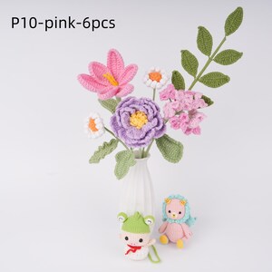 May include: A bouquet of crocheted flowers in pink, purple, and white, arranged in a white vase. The flowers are made of yarn and have a realistic appearance. Two crocheted animal figures, a frog and a pig, are in the foreground.