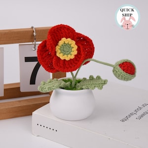 May include: A crocheted poppy flower in a white pot with green leaves. The flower is red with a yellow and green center. The pot is sitting on a white book with the text "Flowers for your favorite..." on the side.