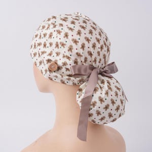 May include: A scrub cap featuring a repeating pattern of brown gingerbread men and small red and green stars on a white background. The cap includes a light brown button on the side and a smooth, light brown ribbon tied into a bow at the back.