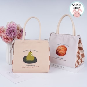 May include: Two beige canvas lunch bags with brown and white checkered accents. One bag features a pear illustration and the text "Plus doux que l'amour" and "DATIOFACTION DE VE Folle joie barmy souvent". The other bag features a pastry illustration and the text "Plus doux que l'amour" and "DATIOFACTION DE VE Folle joie barmy souvent".
