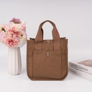 Pode incluir: Uma bolsa tote de lona castanha com duas al&ccedil;as de tecido resistentes e um bolso frontal preso por um bot&atilde;o de press&atilde;o de metal prateado. A bolsa apresenta uma estrutura retangular com costuras refor&ccedil;adas na base das al&ccedil;as.