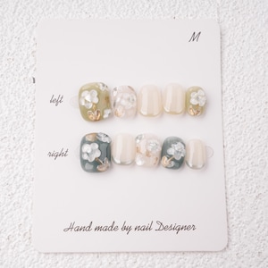 May include: A set of press-on nails in various shades of green, white, and beige, adorned with floral designs and gold accents. The nails are arranged on a white card labeled "M" and "left" and "right". The card reads "Hand made by nail Designer".