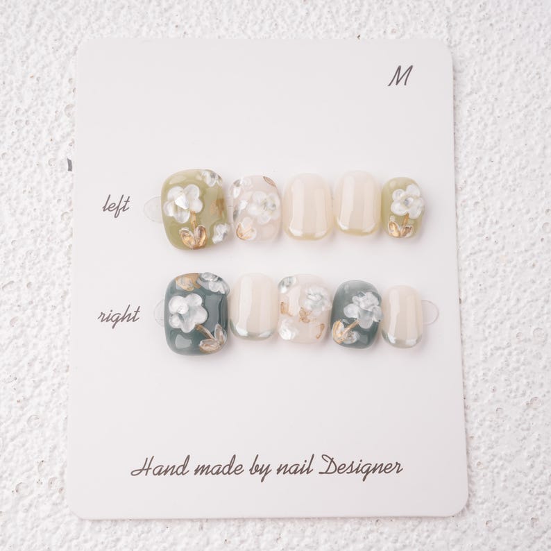 May include: A set of press-on nails in various shades of green, white, and beige, adorned with floral designs and gold accents. The nails are arranged on a white card labeled "M" and "left" and "right". The card reads "Hand made by nail Designer".