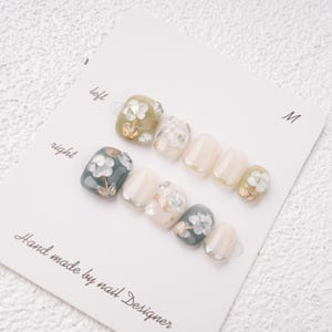 May include: A set of press-on nails with floral designs. The nails feature a mix of colors, including olive green, white, and teal, with gold accents. The card reads "Hand made by nail Designer."