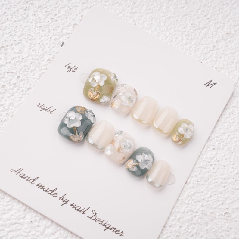 May include: A set of press-on nails with floral designs. The nails feature a mix of colors, including olive green, white, and teal, with gold accents. The card reads "Hand made by nail Designer."