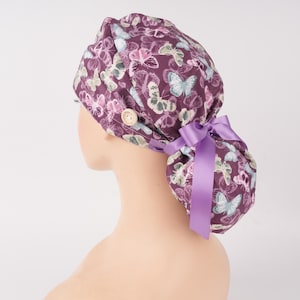 May include: A plum-colored scrub cap featuring a repeating pattern of butterflies in shades of white, light blue, and green. The cap includes a light-colored circular button on the side and a bright lavender ribbon tied into a bow at the back.