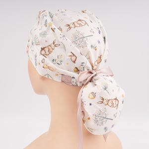 May include: A scrub cap featuring a white background with a repeating pattern of brown rabbits, acorns, and green foliage. The cap includes a gathered back section secured with a light pink ribbon tie and a small wooden button on the side.