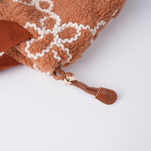 May include: Close-up of a brown faux fur tote bag with a white embroidered pattern. The bag has a brown leather drawstring with a gold metal toggle.