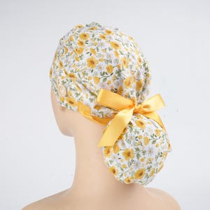 May include: A scrub cap featuring a white background with a yellow and green floral pattern. The cap includes a side button for mask attachment and a bright yellow satin ribbon tied into a bow at the back to secure the gathered fabric pouch.