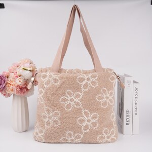 May include: A beige tote bag with a fuzzy texture and white embroidered flower designs. The bag has two long straps.