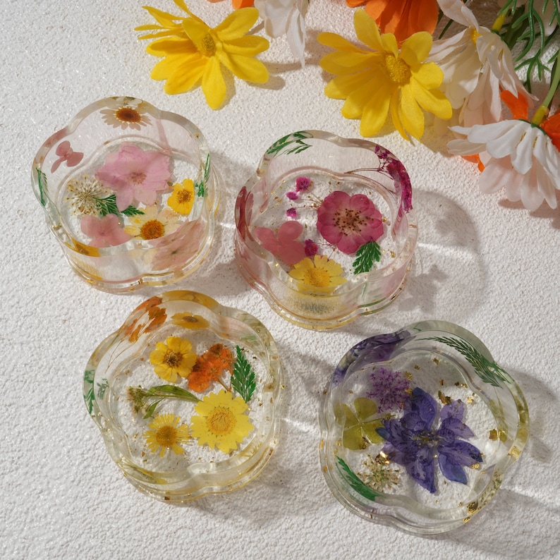 K&ouml;nnte beinhalten: Vier transparente Harz-Untersetzer mit gepressten Blumen und goldenen Blatt-Akzenten. Die Untersetzer sind rund und haben einen gewellten Rand. Die Blumen sind in verschiedenen Farben, darunter rosa, lila, gelb und wei&szlig;.