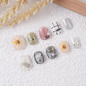 May include: A set of ten press-on nails featuring various designs including floral patterns, plaid, glitter, and pearl embellishments. The collection includes shades of pink, green, white, and gray with metallic accents and small beads.