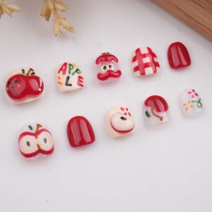 May include: Set of ten red and white nail art tips featuring apple designs. The designs include whole apples, apple slices, and the word "APPLE".