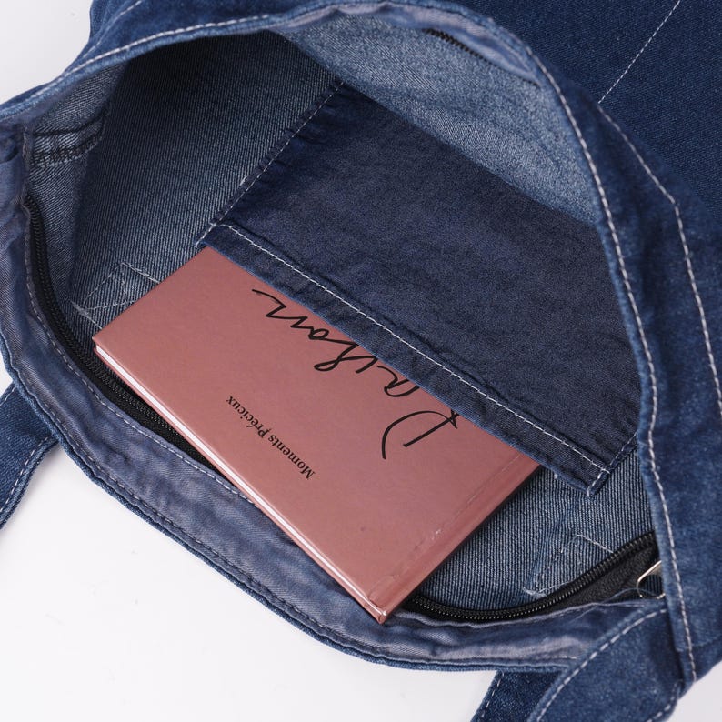 Peut inclure: Un sac fourre-tout en jean bleu avec des coutures blanches et une poche zipp&eacute;e. &Agrave; l'int&eacute;rieur de la poche se trouve un livre intitul&eacute; "Daison" et les mots "Moments Pr&eacute;cieux" imprim&eacute;s sur la couverture.