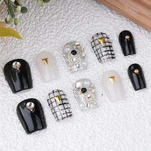 May include: Set of ten false nails with different designs. The nails are black, white, and silver with gold accents. Some nails have rhinestones and others have a checkered pattern.