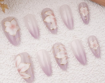 Könnte beinhalten: Set mit zehn falschen Nägeln mit einem hellvioletten Ombre-Finish. Jeder Nagel hat ein zartes weißes Blumendesign mit goldenen Akzenten.