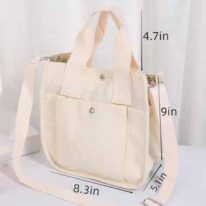 Pode incluir: Uma bolsa tote de lona cor de creme com duas al&ccedil;as superiores e uma al&ccedil;a de ombro remov&iacute;vel. A bolsa possui um bolso frontal com fecho de bot&atilde;o de press&atilde;o prateado. Dimens&otilde;es: 21,1 cm de largura, 22,9 cm de altura, 13 cm de profundidade.