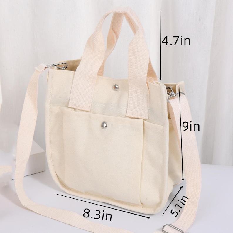 Pode incluir: Uma bolsa tote de lona cor de creme com duas al&ccedil;as superiores e uma al&ccedil;a de ombro remov&iacute;vel. A bolsa possui um bolso frontal com fecho de bot&atilde;o de press&atilde;o prateado. Dimens&otilde;es: 21,1 cm de largura, 22,9 cm de altura, 13 cm de profundidade.