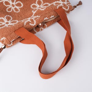 May include: A brown tote bag with white floral embroidery and orange straps. The bag has a zipper closure and a small handle on the top.