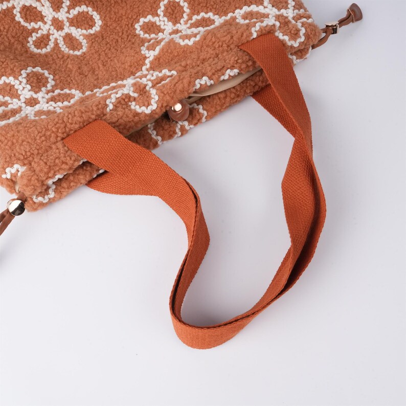 May include: A brown tote bag with white floral embroidery and orange straps. The bag has a zipper closure and a small handle on the top.