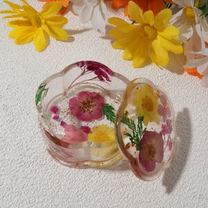K&ouml;nnte beinhalten: Eine transparente Schmuckschatulle aus Harz mit einem gewellten Rand, gef&uuml;llt mit gepressten Blumen und goldenen Flocken. Der Deckel ist ge&ouml;ffnet und zeigt ein &auml;hnliches Design mit rosa und gelben Blumen.