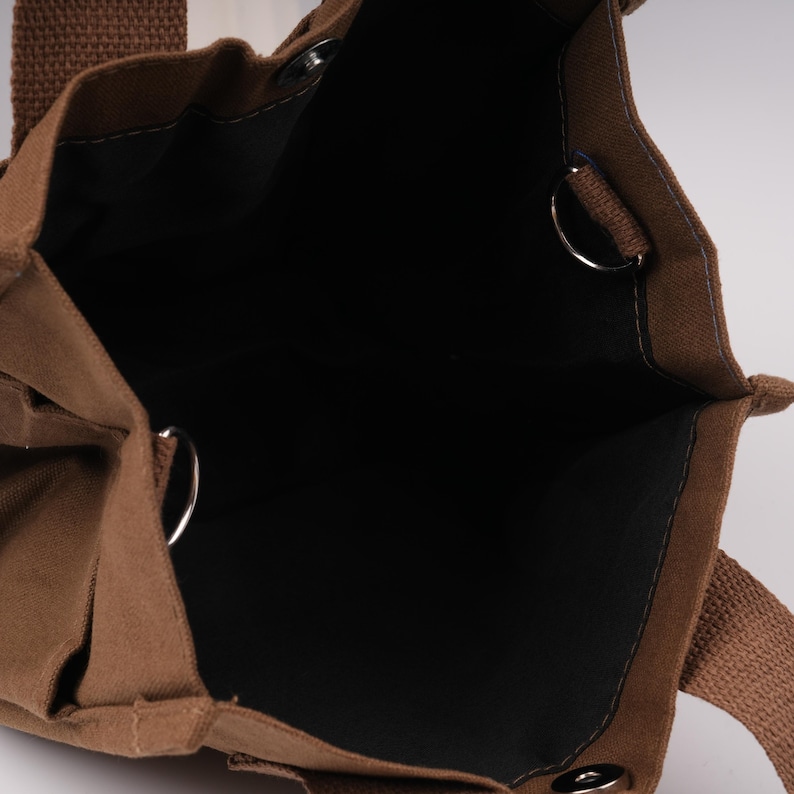 Pode incluir: Uma bolsa tote de lona castanha com forro interior escuro, apresentando duas al&ccedil;as de tecido castanho resistentes, argolas em D de metal prateado fixadas nas laterais internas e um fecho de bot&atilde;o de press&atilde;o prateado na abertura superior.