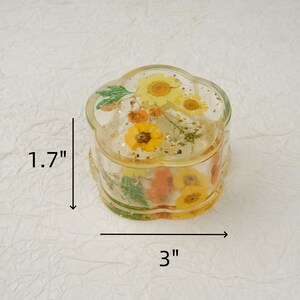 K&ouml;nnte beinhalten: Eine runde, transparente Schmuckdose in Blumenform mit Deckel. Die Dose ist mit gepressten Blumen und Blattgold gef&uuml;llt. Die Dose hat einen Durchmesser von 7,6 cm und eine H&ouml;he von 4,3 cm.