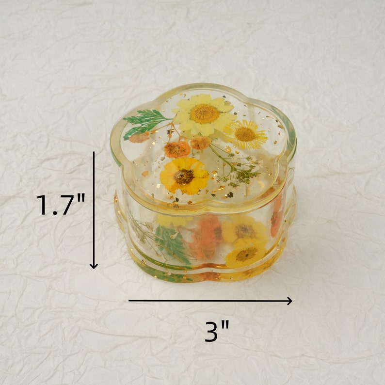 K&ouml;nnte beinhalten: Eine runde, transparente Schmuckdose in Blumenform mit Deckel. Die Dose ist mit gepressten Blumen und Blattgold gef&uuml;llt. Die Dose hat einen Durchmesser von 7,6 cm und eine H&ouml;he von 4,3 cm.