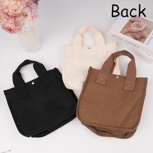 Pode incluir: Tr&ecirc;s pequenas bolsas de lona nas cores preta, creme e marrom. Cada bolsa possui duas al&ccedil;as superiores resistentes, uma silhueta quadrada e um fecho de bot&atilde;o de press&atilde;o de metal prateado no centro da abertura. As bolsas est&atilde;o dispostas juntas.
