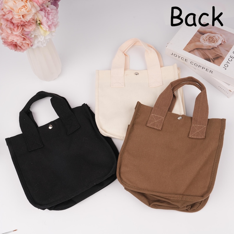 Pode incluir: Tr&ecirc;s pequenas bolsas de lona nas cores preta, creme e marrom. Cada bolsa possui duas al&ccedil;as superiores resistentes, uma silhueta quadrada e um fecho de bot&atilde;o de press&atilde;o de metal prateado no centro da abertura. As bolsas est&atilde;o dispostas juntas.