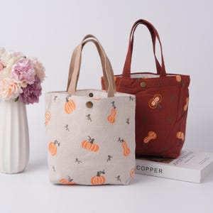May include: Two tote bags. One is beige with orange pumpkin and vine designs, tan handles, and a snap closure. The other is rust-colored with a similar design, a snap closure, and a matching handle. A vase of flowers is on the left.