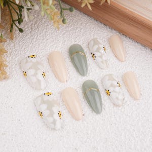 May include: Set of ten false nails with a white and yellow daisy design. The nails are in a variety of shapes and sizes, including almond, oval, and square. Some nails are a light beige color, while others are a light green color with a gold accent.