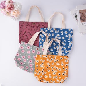 May include: Four floral tote bags with cream-colored handles. The bags feature different floral patterns: a burgundy floral print, a blue daisy print, a gray and pink floral print, and an orange daisy print. Each bag has a snap closure.