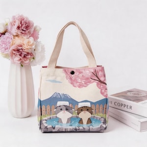 May include: A canvas tote bag featuring a printed illustration of two cats in a hot spring with a mountain and cherry blossom trees in the background. The bag has beige fabric handles and a single black snap button closure at the top center.