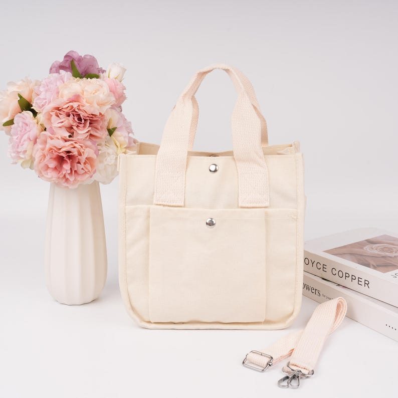Pode incluir: Uma bolsa tote de lona na cor creme com duas al&ccedil;as superiores, um bolso frontal com fecho de bot&atilde;o de press&atilde;o prateado e uma al&ccedil;a de ombro remov&iacute;vel a condizer com clipes de metal prateado. A bolsa tem formato quadrado e fecho de press&atilde;o.