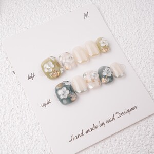 May include: A set of press-on nails with a floral design. The nails are arranged on a white card labeled "left" and "right". The nails feature a variety of colors, including white, green, and gold accents. The text "Hand made by nail Designer" is printed on the card.