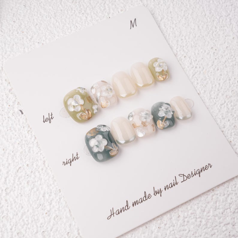 May include: A set of press-on nails with a floral design. The nails are arranged on a white card labeled "left" and "right". The nails feature a variety of colors, including white, green, and gold accents. The text "Hand made by nail Designer" is printed on the card.