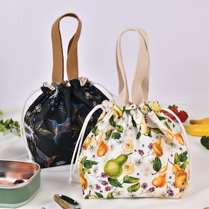 May include: Two drawstring lunch bags. One is black with a floral and hummingbird pattern, and the other is white with a pear and floral design. Both bags have beige handles and white drawstring closures.