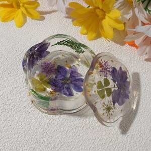 K&ouml;nnte beinhalten: Eine transparente Schmuckschatulle aus Harz mit einem gewellten Deckel. Die Schachtel ist gef&uuml;llt mit gepressten lila Blumen, gr&uuml;nen Bl&auml;ttern und goldenen Flocken.