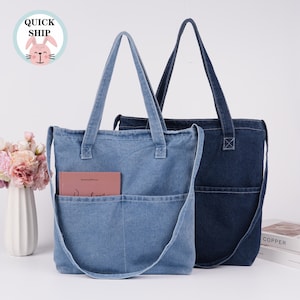 May include: Two denim tote bags, one light blue and one dark blue, with long straps and front pockets. The light blue bag has a book in its pocket. A "QUICK SHIP" sticker with a bunny is in the upper left corner.